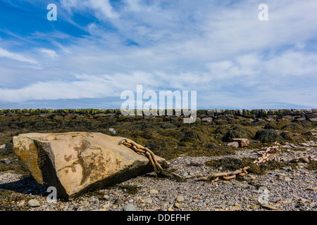 Eine schwere Kette an einem sehr großen Felsbrocken befestigt sitzt am Ufer bei Lubec Maine mit dem steinernen Wellenbrecher im Hintergrund. Stockfoto