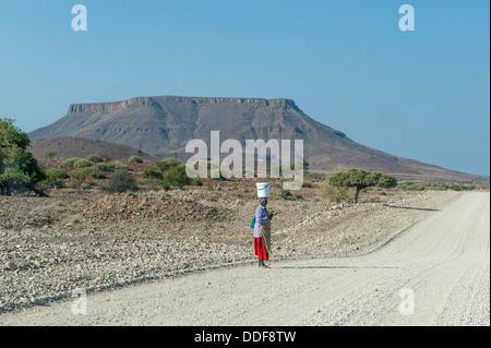 Eine Frau mit einem Eimer auf ihrem Kopf stehend auf einer Schotterstraße in trockene Landschaft, Kunene Region, Namibia Stockfoto