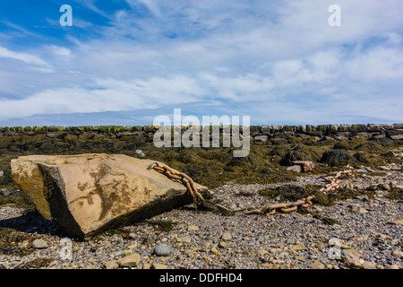 Eine schwere Kette an einem sehr großen Felsbrocken befestigt sitzt am Ufer bei Lubec Maine mit dem steinernen Wellenbrecher im Hintergrund. Stockfoto