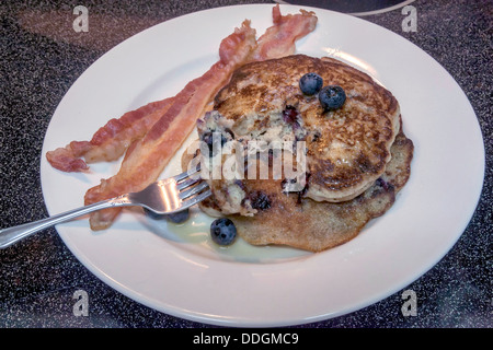 Gabel ruht auf Platte mit zwei großen Blaubeerpfannkuchen garniert mit Heidelbeeren, Ahornsirup und zwei Streifen Speck. Stockfoto