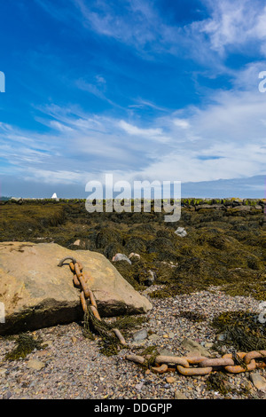 Eine schwere Kette an einem sehr großen Felsbrocken befestigt sitzt am Ufer bei Lubec Maine mit dem steinernen Wellenbrecher im Hintergrund. Stockfoto