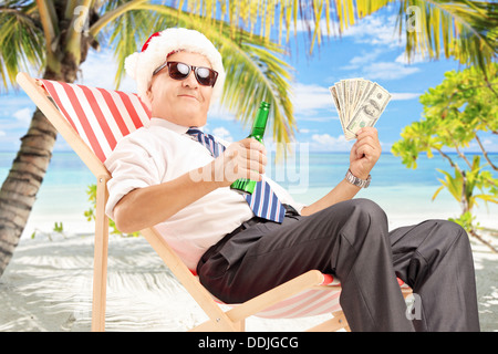 Lächelnd Geschäftsmann mit Weihnachtsmütze auf einem Stuhl sitzend und hält US-Dollar und Bier, an einem Strand in der Nähe des Meeres Stockfoto