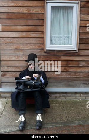 seltsamer Mann mit schwarzem Mantel und Hut halten Koffer mit Kätzchen sitzen auf Bürgersteig in der Nähe von Holzhaus in Regen und Schnee Stockfoto