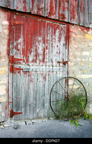 eine rustikale alte Scheunentor mit peeling, rote Farbe, Steinmauern und einer verrosteten Antik Wagenrad Stockfoto