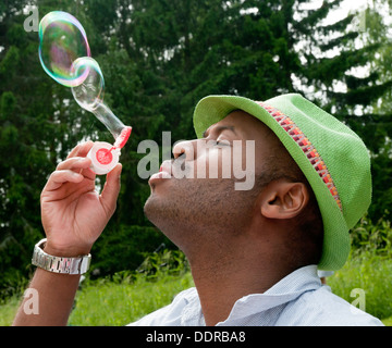 Hübscher Junge afroamerikanischer Mann bläst Seifenblasen in einer losen Businesshemd, grünen Hut mit grünem Rasen und Wald hinter. Stockfoto