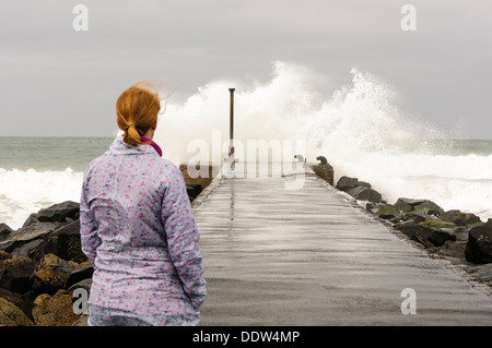 Eine Frau schaut zu, wie eine Welle über das Ende eines Piers in den windigen Bedingungen stürzt ab Stockfoto