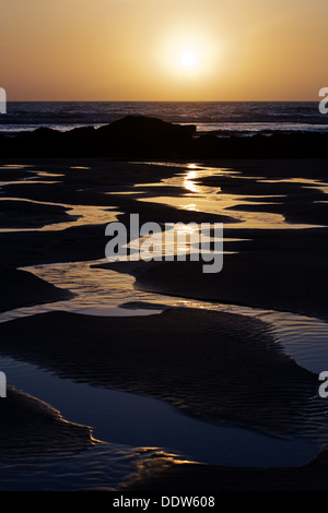 Hell orange untergehende Sonne spiegelt sich in den gewundenen Bächen Wasser am Strand von Porthtowan in Cornwall, Großbritannien Stockfoto