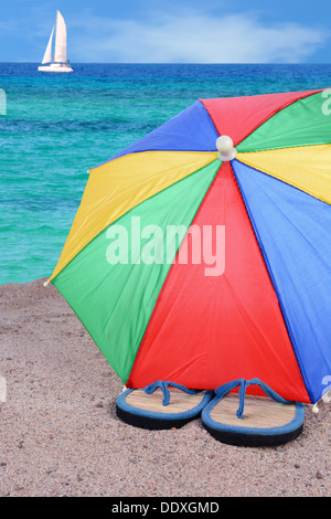 Nahaufnahme eines Paares Flip-Flops teilweise unter einem bunten Sonnenschirm. Ein Meer malerisch mit Segelboot im Hintergrund Stockfoto