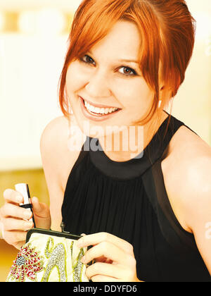 Lächelnde junge elegante Frau in Abendgarderobe, einen Lippenstift aus ihrer Abendtasche Stockfoto