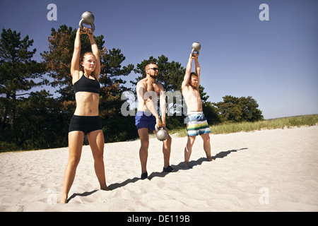 Gruppe von Athleten schwingen eine Wasserkocher Glocke über dem Kopf am Strand. Junge Menschen dabei Crossfit Training an einem heißen Sommertag. Stockfoto