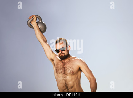 Männlicher Athlet eine Wasserkocher Glocke über seinen Kopf mit einer Hand schwingen. Junge Hipster Mann tut Crossfit Training an einem heißen Sommertag. Stockfoto