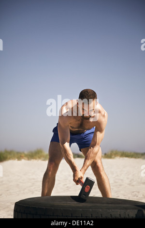 Männlicher Athlet Hämmern LKW-Reifen mit einem Vorschlaghammer beim Crossfit Training am Strand Stockfoto