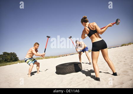 Drei starke Athleten tun Hammer Streik auf einem LKW-Reifen beim Crossfit Training draußen am Strand Stockfoto