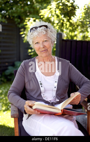 Glücklich Reife Frau sitzend auf einem Stuhl im Garten mit Blick auf die Kamera lächelnde - ältere Frau im freien lesen Buch Stockfoto