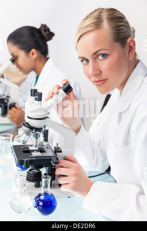 Ein blondes medizinische oder wissenschaftliche Forscherin oder Arzt mit ihrem Mikroskop im Labor mit ihrem asiatischen Kollegen. Stockfoto