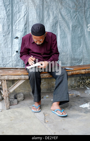 älterer Mann sitzt auf einem klapprigen Bambus Bank am Straßenrand Schreiben von Notizen Kediri Java Indonesien Stockfoto