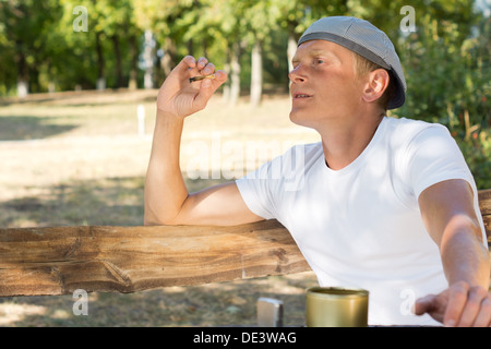 Mann, sitzen, Rauchen eine Zigarette oder Gelenk von Marihuana im Park mit einem kleinen Behälter von Tabak auf dem Tisch vor ihm Stockfoto