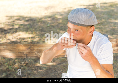 Mann mittleren Alters Rollen eine Zigarette, die Abdichtung des Papiers sitzen auf einer Bank im Park an einem sonnigen Tag des Sommers Stockfoto