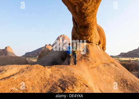 Zwei junge Männer stehen unter einem Bogen Felsen, in der Nähe der Spitzkoppe Granitfelsen, Damaraland, Namibia, Afrika Stockfoto