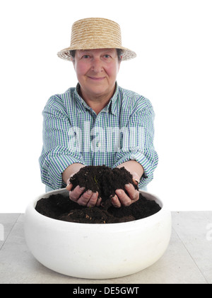 Weibliche senior Gärtner Stroh Hut hält eine Handvoll Erde über weißen Keramiktopf Stockfoto