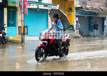 Indischer Mann mit einem Regenschirm auf seinem Motorrad reiten durch nasse Straßen in Puttaparthi, Andhra Pradesh, Indien Stockfoto