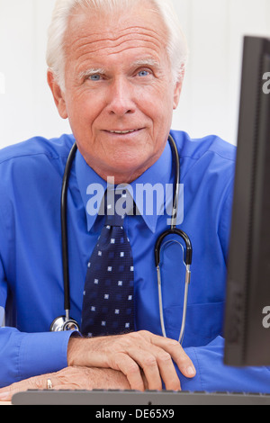 Ein senior männlichen Arzt sitzen an einem Schreibtisch in einem Büro mit Computer und trug ein Hemd, Krawatte und Stethoskop Stockfoto