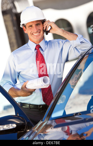 Ein erfolgreicher Geschäftsmann oder Architekten tragen einen weißen harten Hut hält Pläne & reden auf seinem Handy von einem Auto Stockfoto