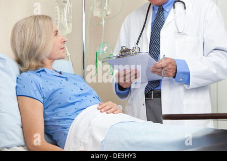 Ältere Frau in einem Krankenhausbett mit Gespräch mit männlichen Arzt mit Stethoskop und Zwischenablage Stockfoto