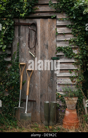 Rustikale alte Gartenhaus bedeckt mit Efeu mit einer Gabel und Spaten stützte sich auf die Tür. Stockfoto