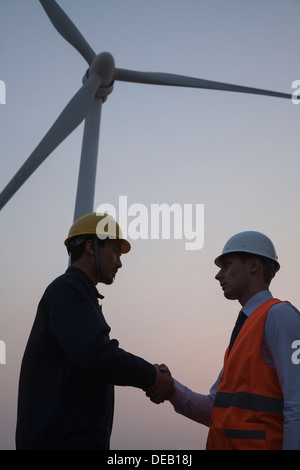 Zwei junge männliche Ingenieure stehen neben einer Windkraftanlage bei Sonnenuntergang und Händeschütteln Stockfoto