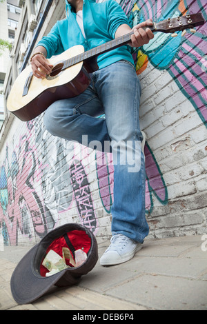 Young Straßenmusikant Gitarre zu spielen und als Straßenmusikant Geld vor einer Wand mit graffiti Stockfoto