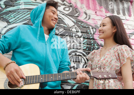 Junge lächelnde Straßenmusiker stützte sich auf Wand mit Graffiti-Zeichnungen, Gitarre zu spielen und flirten mit einer jungen Frau in einem Kleid Stockfoto