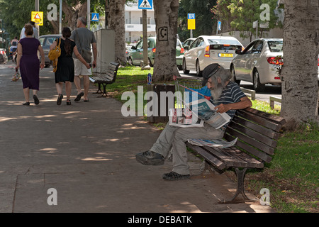 Ein Obdachloser Mann liest eine Zeitung auf einer Bank im Rothschild Boulevard, während Menschen vorbeiziehen. Tel Aviv, Israel Stockfoto