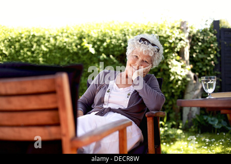 Alte Frau auf Stuhl Blick in die Kamera. Senior weiblich entspannend im Garten genießen ihren Ruhestand Tage Stockfoto