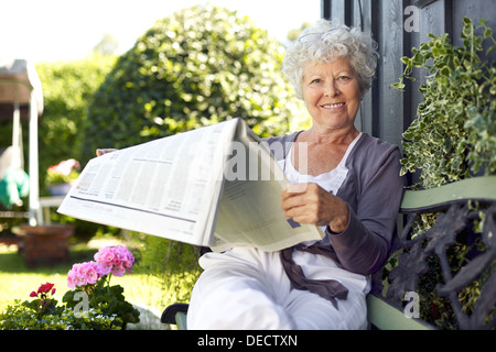 Entspannte senior Frau sitzen auf einer Bank im Garten Lesen einer Zeitung Blick in die Kamera und lächelnd Stockfoto