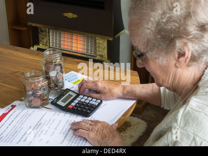 Ältere Dame in ihren neunziger Jahren, die Gas-, Elektro-, Stromrechnung mit Taschenrechner und Geldgefäße auf dem Tisch betrachtet. Gasfeuer im Hintergrund Stockfoto