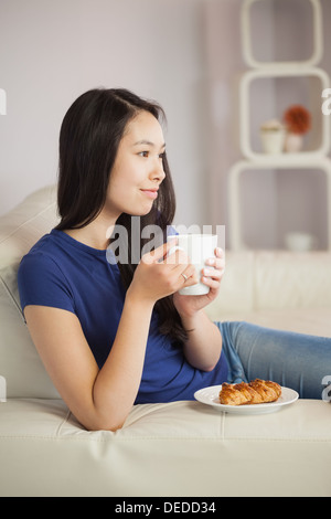 Junge asiatische Frau sitzt auf dem Sofa mit Kaffee withpastry Stockfoto