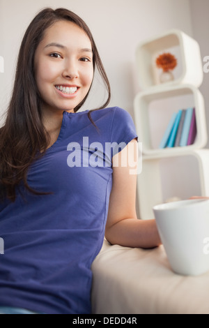 Glückliche junge asiatische Frau sitzen auf der Couch halten Becher Stockfoto