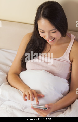Fröhliche junge asiatische Frau sitzen im Bett SMS auf ihrem smartphone Stockfoto