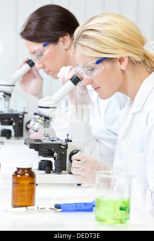 blonden medizinische oder wissenschaftliche Forscherin oder Arzt mit ihrem Mikroskop im Labor mit ihrer Kollegin Stockfoto