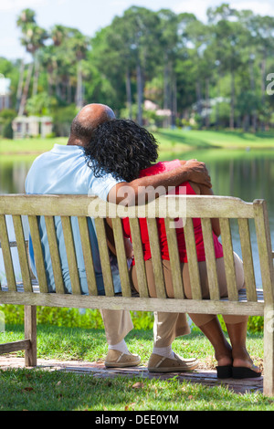 Rückansicht des einen romantischen senior afroamerikanische Brautpaar sitzt auf einer Parkbank, umarmen, Blick auf einen See Stockfoto