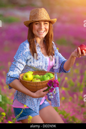 Porträt von niedlichen fröhliches Mädchen mit Apfelkorb auf rosa Blumen Feld tragen, Strohhut, Ernte, Herbst Natur Stockfoto