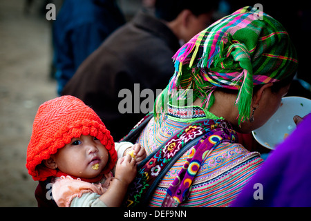 Ein Baby Flower Hmong blickt auf, während ihre Mutter Mittagessen am Sonntag Morgen Markt in Bac Ha, Provinz Lao Cai, Vietnam isst. Stockfoto