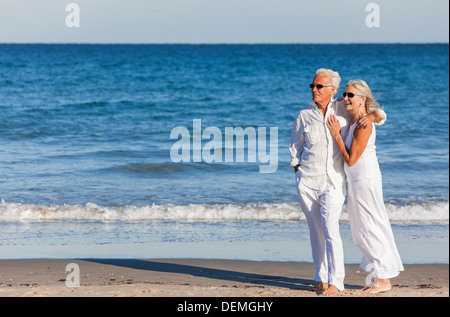 Glücklich senior Mann und Frau Paar zusammen auf dem Seeweg auf einem einsamen tropischen Strand mit strahlend blauem Himmel umarmen Stockfoto