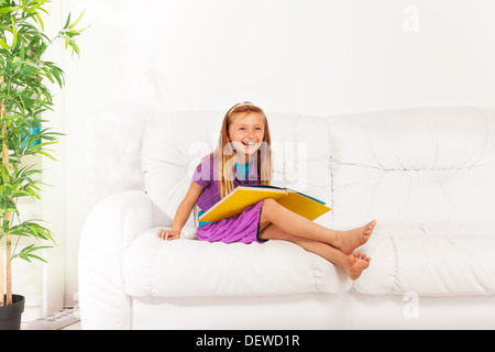 Sehr glücklich und klug lachende blonde langhaarige Mädchen sitzen auf der weißen Couch mit großen gelben Buch Stockfoto