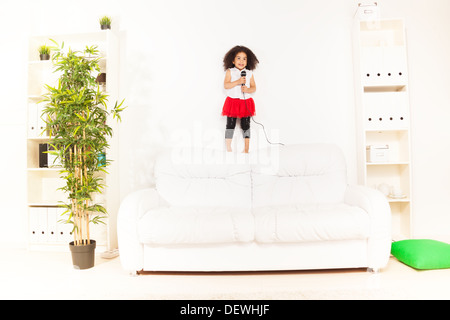 Niedliche kleine schwarzen afrikanischen drei Jahre alten Mädchen mit Mikrofon durchführen stehen auf dem Sofa im Wohnzimmer Stockfoto