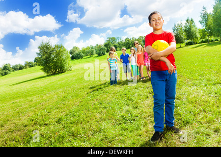 Porträt eines lächelnden asiatischen Jungen mit Volleyball Ball stehend im Park mit seinem Team auf Hintergrund Stockfoto