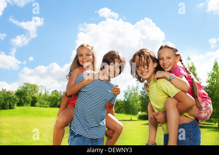 Zwei Paar, Mädchen reitet auf des Jungen zurück, beide glücklich und lächelnd, im Park an einem sonnigen Tag im Park mit blauem Himmel und wenig Wolken Stockfoto