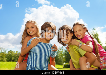 Zwei Kinder, Mädchen reitet auf des Jungen zurück, beide glücklich und lächelnd, im Park an einem sonnigen Tag im Park mit blauem Himmel und wenig Wolken Stockfoto