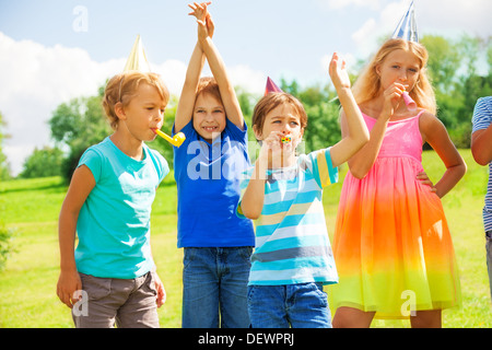 Gruppe von vier Kinder draußen auf einer Geburtstagsfeier weht Krachmachern Hörner und Pfeifen verdreht Stockfoto